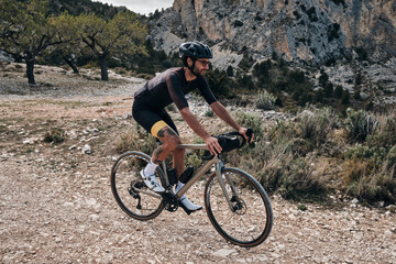 Naklejka na ściany i meble Man riding gravel bike on gravel road in mountains with scenic view.Professional cyclist practicing on gravel road.Male cyclist wearing black cycling kit and helmet.Cycle camp in Calpe, Alicante,Spain