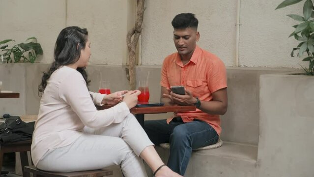Full shot of Indian man holding smartphone and discussing something on screen with female colleague while sitting in cafe and having drinks