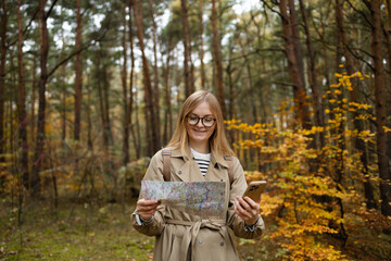 Fototapeta na wymiar Beautiful blonde woman holding mobile phone using map to navigate in autumn forest. Happy tourist trying to find best way. Active lifestyle, travel concept. High quality photo