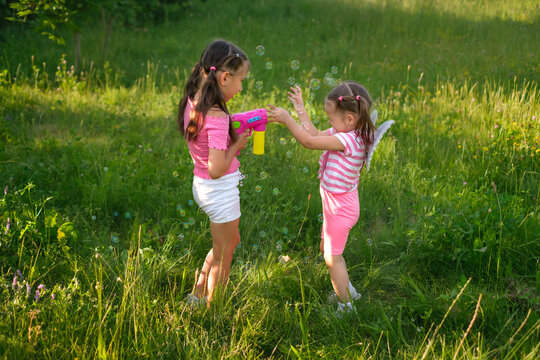 A kid shoots air bubbles from an automatic pistol at a little girl covering her face with her hands. Fairy wings behind the child's back