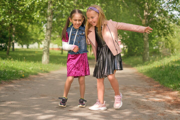 A cute beautiful girl with a broken arm and a cast walks laughing with a friend in the park