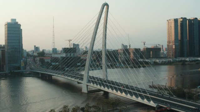Aerial shot of cable-stayed road bridge with traffic over the Saigon river in Ho Chi Minh city, downtown in the background. Golden hour time, sun reflecting in glass buildings and water surface