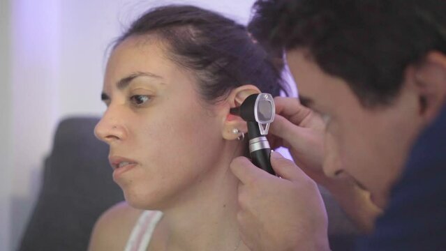 Young Latin woman having her ears checked by a doctor with an otoscope while she tells him about the symptoms she has been having.