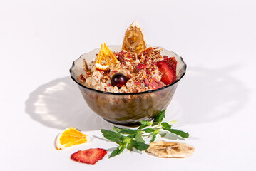 Healthy breakfast. Oatmeal with pieces of fruits and berries in a transparent bowl isolated on a white background. Scattered mint, dried banana, orange and strawberry slice. Healthy snacks.