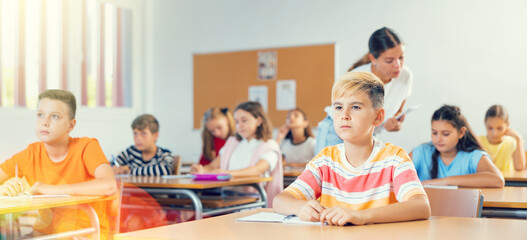 Diligent elementary school student tween boy studying with classmates, making notes of teacher lecture