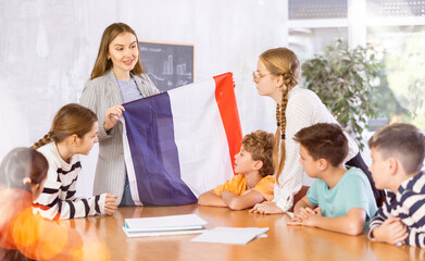 Decent teacher showing France flag to group of preteen schoolchildren in classroom during lesson