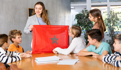 Fototapeta na wymiar Group of preteen schoolchildren attentively watching teacher describing Morocco flag in schoolroom