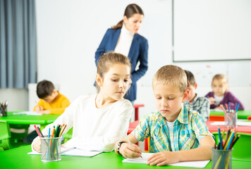 Portrait of diligent preteen students writing exercises at lesson with young female teacher in primary school