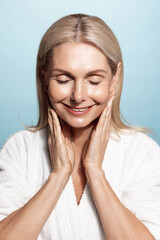 Aging beauty. Vertical portrait of middle aged, elderly woman, 50 years, touches her smooth, nourished skin, applies facial cream, moisturizer, smiling pleased, wears bathrobe, blue background