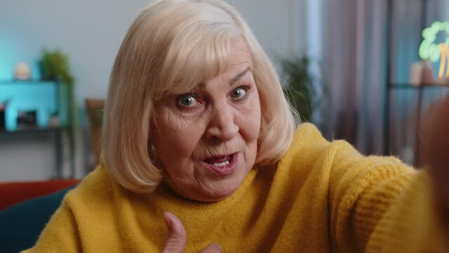 POV of senior grandmother woman making online selfie video call talking waving hand looking at camera at home apartment indoors. Communication, meeting. Elderly blogger making smartphone conversation