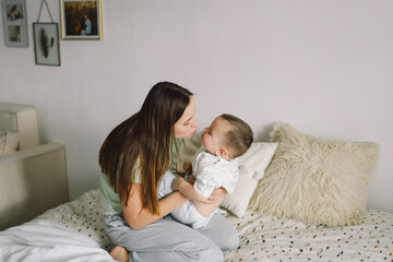 Home portrait of a baby boy with mother on the bed relaxing and playing in the bed at the weekend together, lazy morning, warm and cozy scene. Mom holding and kissing her child. Mother day concept