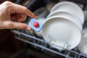 Close up of woman hand filling dishwasher tablet into open automatic stainless built-in dishwasher machine with dirty white dish inside in modern home kitchen.Household, housekeeping domestic life. - obrazy, fototapety, plakaty