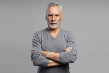 Portrait of serious senior man wearing gray sweater holding arms crossed, looking at camera isolated on gray background