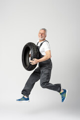 Handsome happy senior man holding and jumping with seasonal tire, wearing overalls isolated on gray background. Shopping, sale, advertisement concept