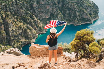 Young woman standing on a rock cliff and waving the US flag while looking at sea beneath. Girl...