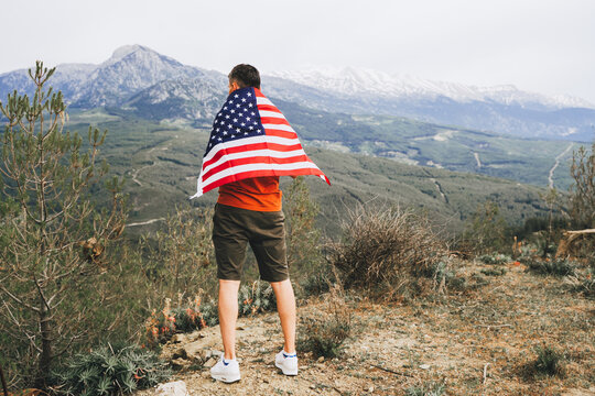 Young man standing on rock cliff with the US flag on his back while looking at mountains in background. Male traveller wearing American flag standing on mountain top. 4 fourth July Independence Day. - Powered by Adobe