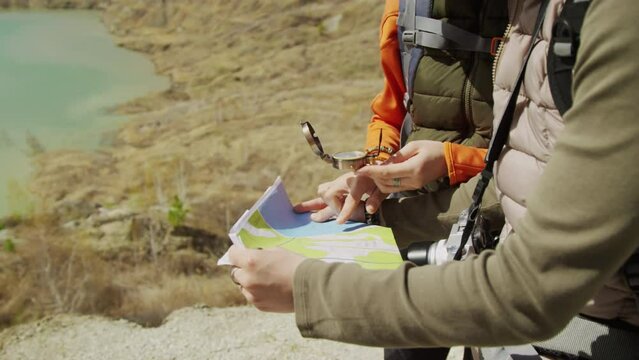 Midsection shot of sunlit map and female hands of two different people, pointing at it, one hand holding compass, wind blowing map. Barren landscape, lake in background
