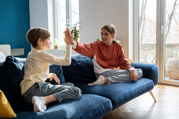 Happy children siblings giving high five to each other while playing together at home, older sister spending time with younger brother, sitting on sofa in living room and having fun