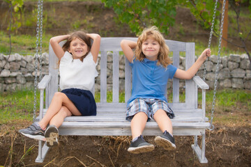 Cute little children playing outdoors. Portrait of two happy young kids swinging in garden wooden swing at the summer park.