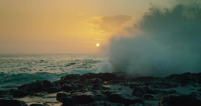 Dim yellow sun on the horizont over the ocean. Sunset orange sky with light reflection on sea water. Huge wave breaks on the rocks and splashes fly into the camera.
