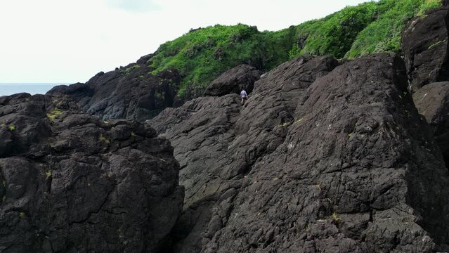 Traveler walks on weathered black lava rocks by blue ocean water and green trees