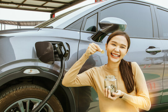 Portrait Young Asian Woman Driving An EV Electric Vehicle During Charging, Smiling Good Mood, Showing Coins In A Glass Jar : Concept Of Using Electric Cars To Save Costs And Have Savings In The Jar.