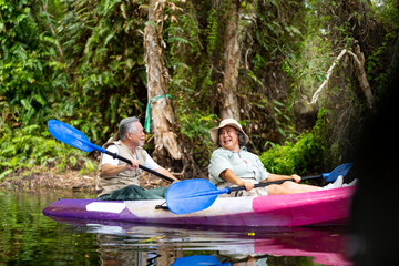 Asian senior couple kayaking together in the lake at mangrove forest on summer vacation. Retired elderly people man and woman have fun outdoor lifestyle travel nature and rowing a boat in the river. - obrazy, fototapety, plakaty