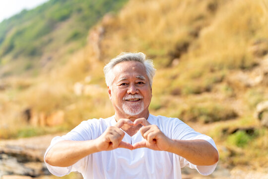 Portrait of Happy Asian senior man showing hands heart shape during walking at tropical beach at summer sunset. Retired elderly people enjoy outdoor lifestyle travel nature ocean on holiday vacation.