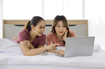 Fototapeta premium Two Asian women spending vacation time in bed, search for investment information on the internet through laptop computer.