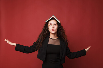 fashion female portrait. Girl, curly girl with black hair in a strict business suit on a red, pink background. High quality photo. Female student rejoices, happy and thoughtful with a book on her head