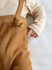 a little baby's hand in a beige bodysuit, and brown pants with suspenders on a white background
