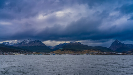A picturesque snow-capped mountain range against the background of the sky and dark rain clouds. City houses on the shore of the strait. Beagle Channel. Argentina. Ushuaia