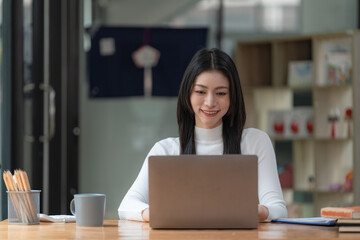 Happy young Asian businesswoman sitting at her workplace in the office. Young woman working at laptop in the office.
