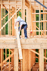 Carpenter building wooden-framed house. Man lifting up large beam, dressed in work clothes and helmet. Concept of contemporary and eco-friendly construction.