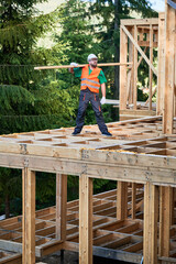 Carpenter constructing wooden-framed house near the woods. Man holds large beam on his shoulder and is dressed in work clothes and helmet. Modern concept of environmentally-conscious building.