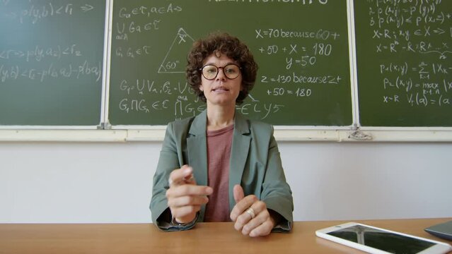 Female Math teacher sitting at desk against blackboard with formulas and equations written on it greeting students and starting online class