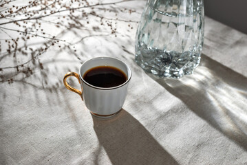 Aesthetic minimalist morning concept, cup of coffee, glass vase and floral sunlight shadows on...