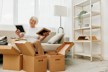 Fototapeta premium elderly woman sits on a sofa at home with boxes. collecting things with memories albums with photos and photo frames moving to a new place cleaning things and a happy smile. Lifestyle retirement.