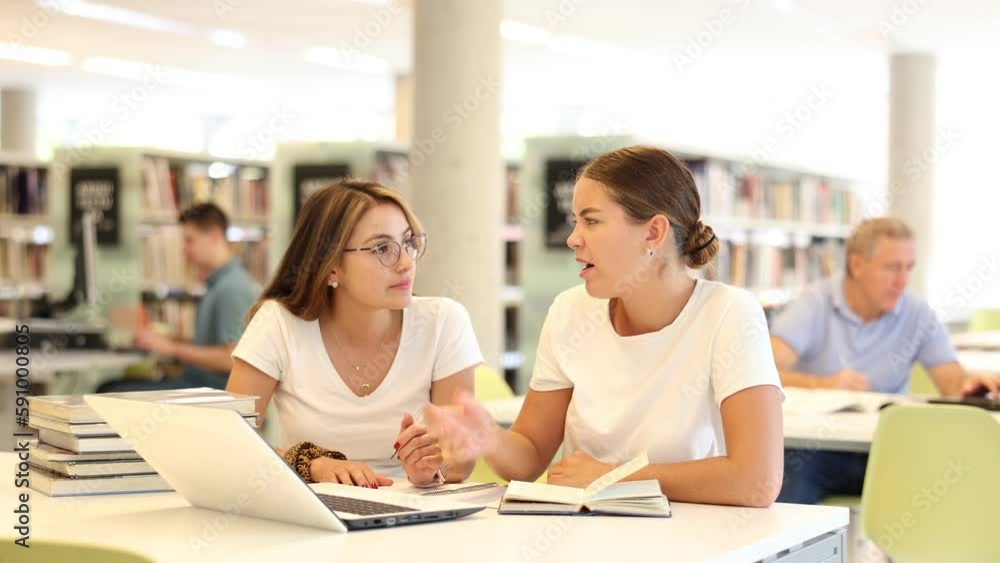 Poster two positive ladies wearing casual clothes behave loudly while studying in the library and using com