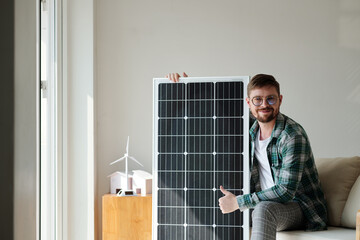 Happy young man sitting next to solar panel and showing thumbs-up