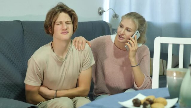 Teenage boy suffering from abdominal pain sitting on sofa at home, holding his stomach and curving through spasms while concerned mother speaking on phone with doctor