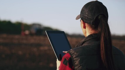 Farmer woman with tablet working in field. Concept of technology in agriculture. Harvest season in an organic farm on a bright sunny day. Tractor at work.