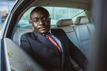 A dark-skinned businessman in a suit in the back seat of a luxury car looks out the window. Premium confidence and comfort