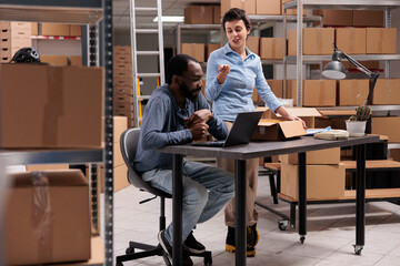 Fashion factory workers analyzing client shipping data on laptop computer after putting clothes order in carton box, discussing packages transportation. DIverse employee working in warehouse.