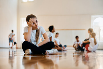 Sad elementary student sits away from her classmates and teacher during PE class at school.