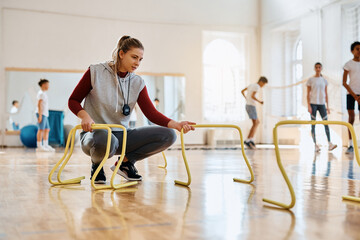 Female coach preparing hurdles for PE class with elementary students at school gym. - Powered by Adobe