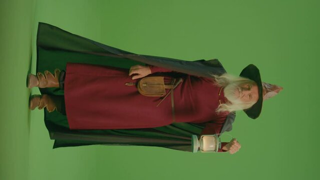 Vertical View.Green Screen. A Portrait of a Smiling Wizard in a Magic Hat and Cloak Walks With a Lamp, Looks Into the Distance and Calls Someone to Him.Magic Forest: a Mysterious and Mysterious World.