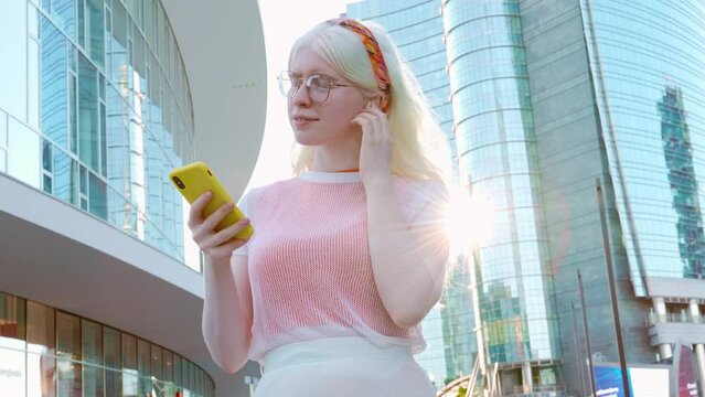 Pretty albino gen z teen girl holding smartphone device wearing earbuds using apps on cell phone listening mobile music looking at smartphone standing on city street lit with sunlight outdoors.