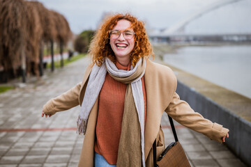 Happy ginger woman with freckles and curly hair is walking on the quay after work. Smiling woman outdoor.
