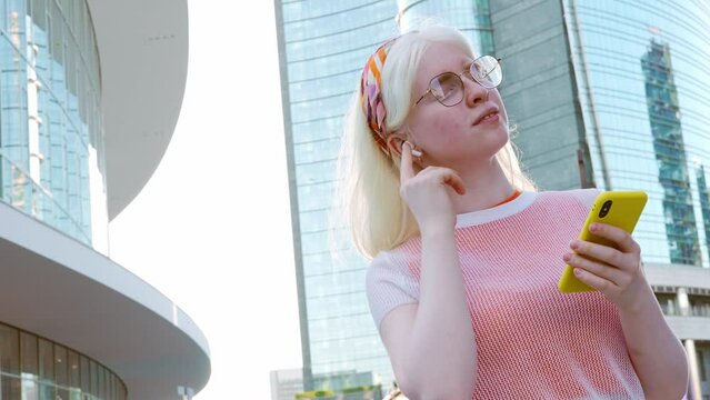 Pretty albino gen z teen girl holding smartphone device wearing earbuds using apps on cell phone listening mobile music looking at smartphone standing on city street lit with sunlight outdoors.